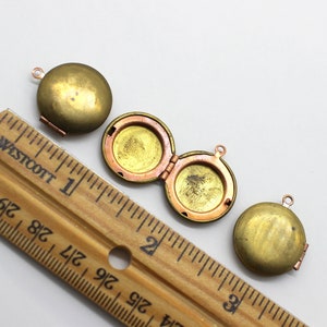 Puede incluir: Tres colgantes de medall&oacute;n de lat&oacute;n antiguo con una abertura con bisagras. Los medallones tienen un di&aacute;metro de aproximadamente 2,5 cm y se muestran junto a una regla.