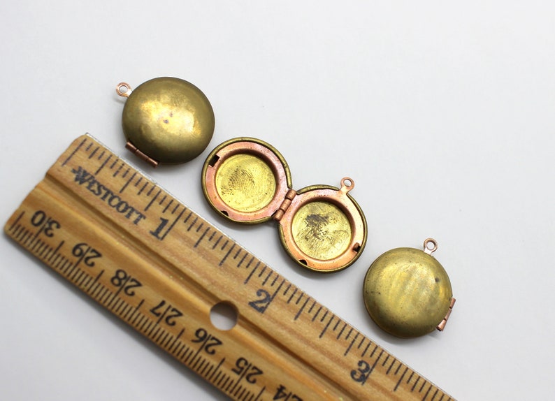 Puede incluir: Tres colgantes de medall&oacute;n de lat&oacute;n antiguo con una abertura con bisagras. Los medallones tienen un di&aacute;metro de aproximadamente 2,5 cm y se muestran junto a una regla.
