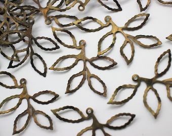 May include: Close-up of several bronze-colored leaf charms scattered on a white surface. Each charm features three oval-shaped leaves connected to a stem with a small loop at the top. The charms have a textured, aged appearance.