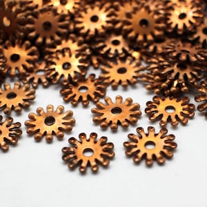 May include: A collection of small, copper-colored metal flower-shaped components. Each piece has a central hole and multiple petal-like projections around the edge. The components are scattered on a white surface.