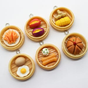 Puede incluir: Encantos de comida en miniatura con varios platos de dim sum, incluyendo maíz, batatas y un bollo al vapor. Cada encanto está en una cesta de vapor de bambú con un lazo plateado.