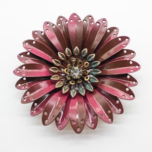 May include: A large flower brooch with layered burgundy and pink petals. The centre has green and gold accents, with a clear crystal. This brooch is designed as a fashion accessory. The brooch is a statement piece.