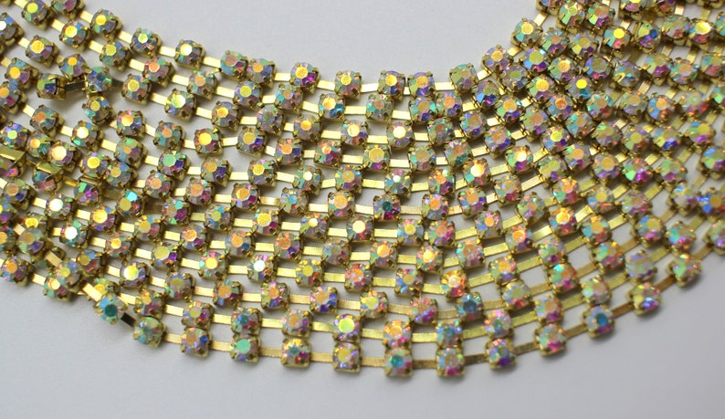 May include: A gold chain with iridescent rhinestones. The chain is made of small, square-shaped rhinestones that are set in a gold metal frame. The rhinestones are arranged in rows and are connected by small gold metal links.