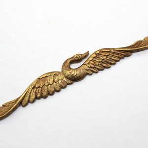 May include: A gold-colored metal swan with outstretched wings. The swan is in a flying position with its head turned to the side. The wings are detailed with feathers.