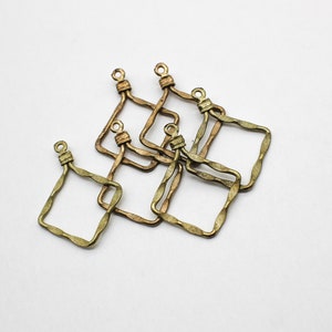 May include: A collection of five antique brass-toned metal charms, each featuring a square frame with a wavy, textured edge and a small circular loop at the top for attachment. The metal has a darkened, weathered finish with visible metallic luster.