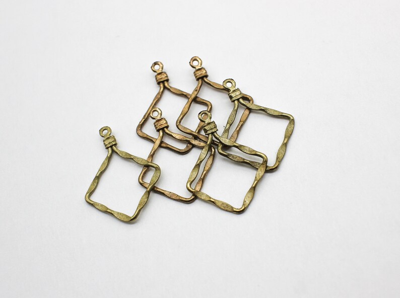 May include: A collection of five antique brass-toned metal charms, each featuring a square frame with a wavy, textured edge and a small circular loop at the top for attachment. The metal has a darkened, weathered finish with visible metallic luster.