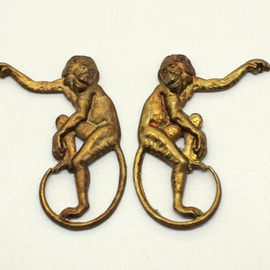 May include: Two gold-colored metal monkey figurines. Each monkey is sitting on its tail, with one arm raised and the other holding its leg.