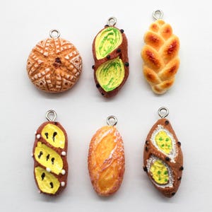 Puede incluir: Una colección de dijes de comida en miniatura, que incluyen un pan redondo y texturizado, una trenza y varios panes ovalados con diferentes ingredientes. Cada dije tiene un lazo plateado para sujetar a joyas o manualidades.