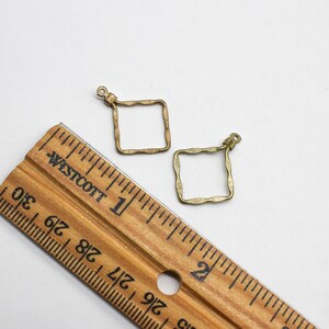 May include: Two antique brass-colored square charms with a wavy, textured border and a small loop at the top for attachment. A wooden Westcott ruler is positioned alongside the charms, showing they measure approximately 0.75 inches in width.