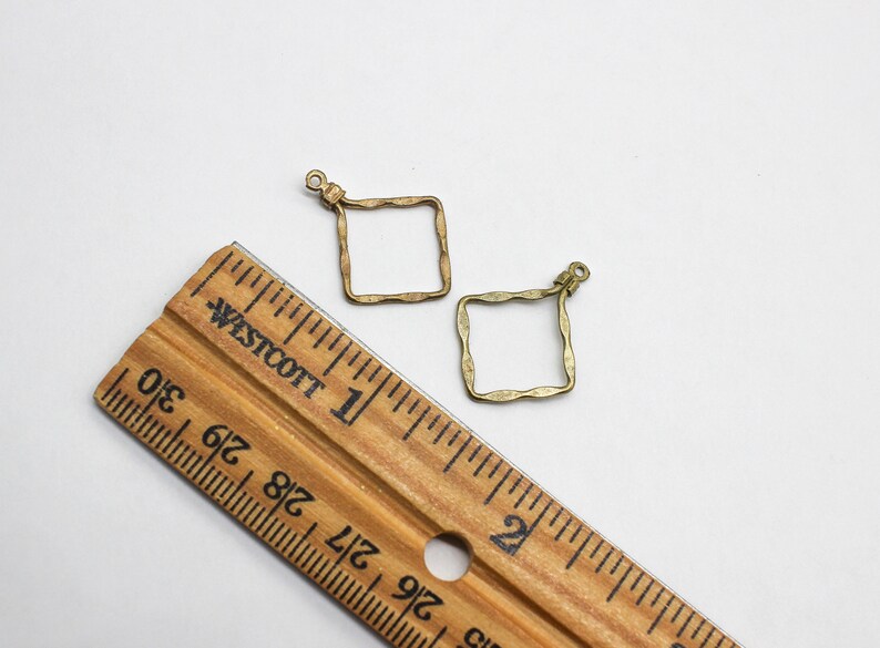 May include: Two antique brass-colored square charms with a wavy, textured border and a small loop at the top for attachment. A wooden Westcott ruler is positioned alongside the charms, showing they measure approximately 0.75 inches in width.