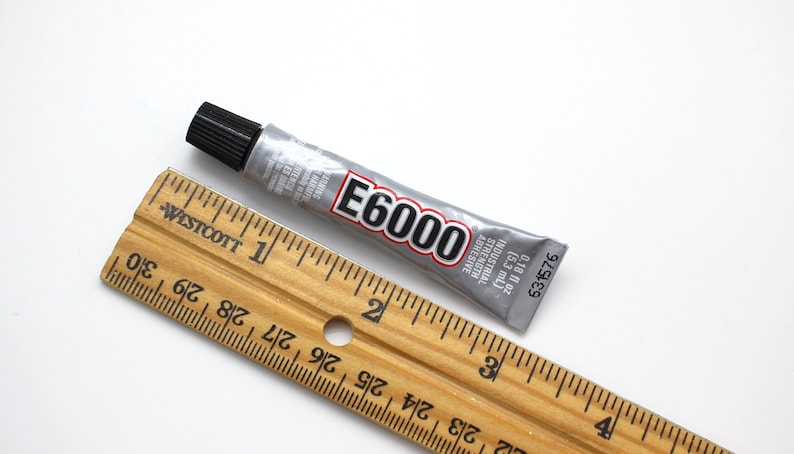 May include: A silver tube of E6000 industrial strength adhesive with a black cap. The tube is 2 inches long and is next to a wooden ruler.