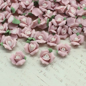 May include: A collection of small, light pink rose-shaped embellishments with white centers and green leaves. These decorative floral accents are scattered across a textured surface, ideal for crafts and DIY projects.