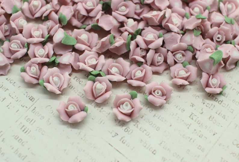 May include: A collection of small, light pink rose-shaped embellishments with white centers and green leaves. These decorative floral accents are scattered across a textured surface, ideal for crafts and DIY projects.