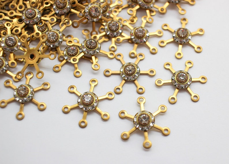 May include: Gold-toned metal snowflake charms with clear rhinestone centers. The charms have six points and are scattered on a white background.