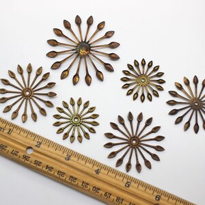 May include: Five gold-toned metal embellishments with a starburst design. Each embellishment has a central circle with radiating points. The embellishments are arranged on a white surface with a wooden ruler for scale.