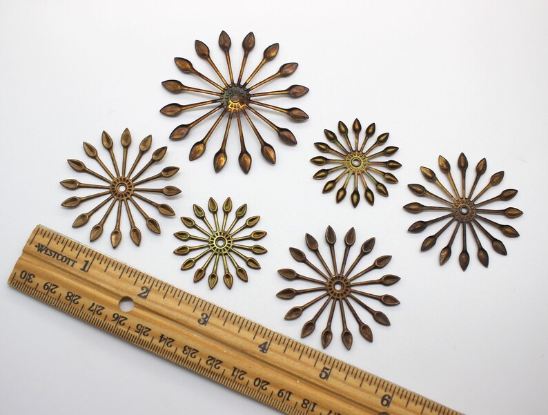 May include: Five gold-toned metal embellishments with a starburst design. Each embellishment has a central circle with radiating points. The embellishments are arranged on a white surface with a wooden ruler for scale.