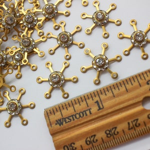 May include: A close-up of a pile of gold-toned metal snowflake charms with clear rhinestones in the center. A ruler is in the background, showing the charms are about 1 inch in diameter.