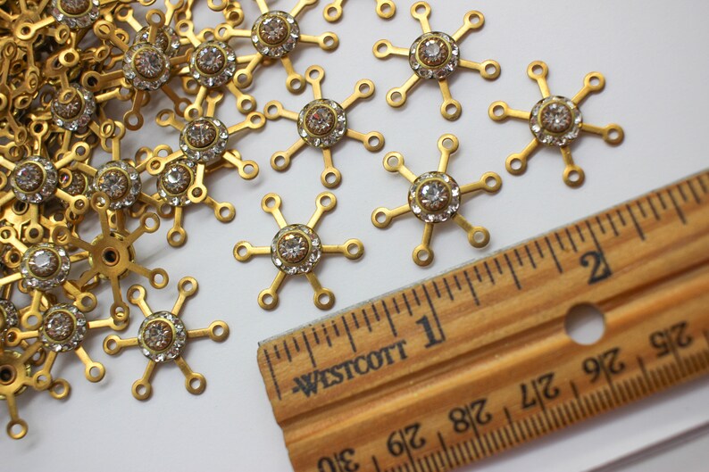 May include: A close-up of a pile of gold-toned metal snowflake charms with clear rhinestones in the center. A ruler is in the background, showing the charms are about 1 inch in diameter.