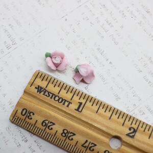 May include: Two small, light pink rose flower beads with green leaves. A wooden ruler with inch markings is in the foreground. The background is a vintage paper with printed text.