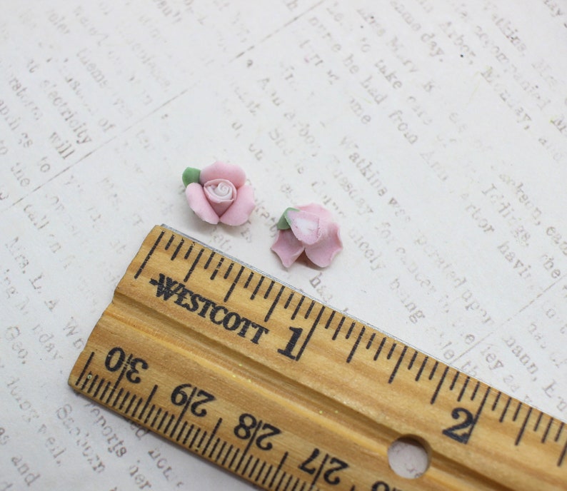 May include: Two small, light pink rose flower beads with green leaves. A wooden ruler with inch markings is in the foreground. The background is a vintage paper with printed text.