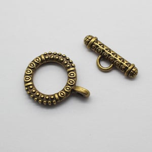 May include: Two antique gold-toned metal clasps for jewelry making. One clasp is a round ring with a decorative pattern. The other clasp is a toggle with a decorative pattern.