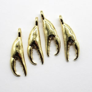May include: Four gold-colored crab claw charms. Each charm has a curved, claw-like design with a loop at the top for attaching to jewelry. The charms are displayed on a white background.