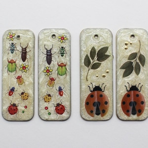 Könnte beinhalten: Vier rechteckige, cremefarbene Anhänger mit Insekten- und Blattmotiven. Die Anhänger zeigen verschiedene bunte Käfer, Marienkäfer und Blätter. Jeder Anhänger hat oben ein kleines Loch zum Aufhängen.
