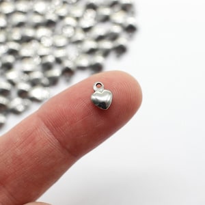 May include: A small, shiny silver heart charm with a loop at the top, held between fingers. The charm is smooth and rounded, with a polished finish. A scattering of similar charms is visible in the background.