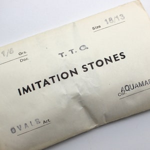 May include: A white paper bag with black text that reads "1/8 Grs. Doz. T.T.C. Size 18/13 IMITATION STONES OVALS Art. Col. AQUAMARINE"