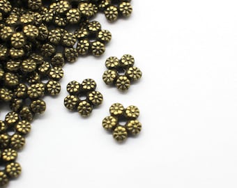 May include: Close-up of antique brass-colored flower-shaped beads. The beads are clustered together on a white background, with several individual beads scattered around. These small, detailed beads are ideal for jewelry making and crafting.