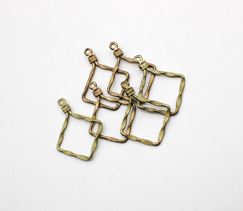 May include: A collection of five square-shaped metal jewelry charms with a textured, wavy edge. Each charm features a small loop at the top for attachment, finished in a muted, antique brass tone with a slightly weathered, metallic appearance.