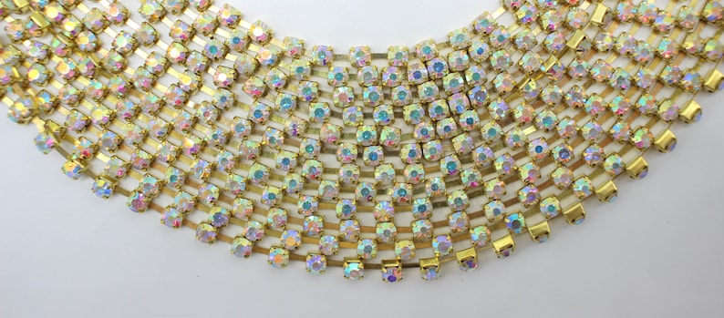 May include: A gold chain with iridescent rhinestones. The chain is made of small, square-shaped rhinestones that are set in a gold metal setting. The chain is about 10 inches long and is perfect for adding a touch of sparkle to any project.