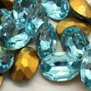 May include: A collection of oval-shaped turquoise blue rhinestones with a faceted design. The rhinestones are scattered on a white surface with a few gold-colored metal pieces.
