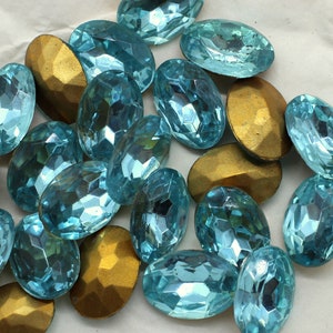 May include: A collection of oval-shaped turquoise blue glass rhinestones with a faceted finish. The rhinestones are scattered on a white surface.
