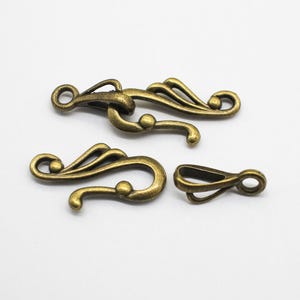 May include: Four antique brass-colored clasps and a clasp component are arranged on a white surface. The clasps have an ornate, flowing design with curved lines and circular accents. The components are likely for jewelry or decorative purposes.
