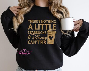 Può includere: Felpa nera con testo in glitter dorato che recita "There's nothing a little Starbucks & Disney can't fix". Il testo è circondato da un contorno in glitter dorato. La felpa è indossata da una persona che tiene una tazza bianca.