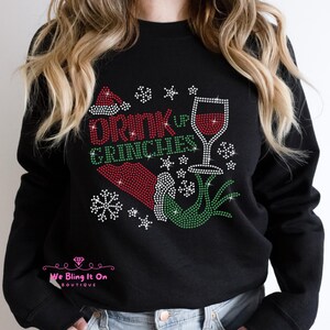 Op de afbeelding: Zwart sweatshirt met een groen en rood strass-ontwerp met de tekst "Drink Up Grinches" met een glas wijn en een kerstmanmuts.