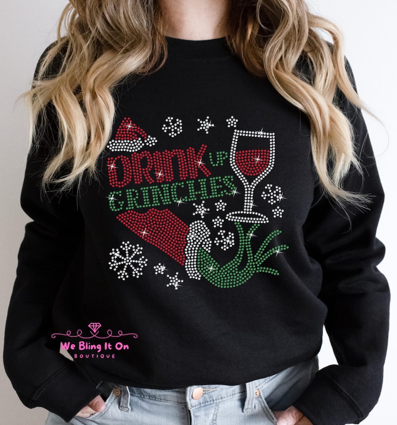 Op de afbeelding: Zwart sweatshirt met een groen en rood strass-ontwerp met de tekst "Drink Up Grinches" met een glas wijn en een kerstmanmuts.