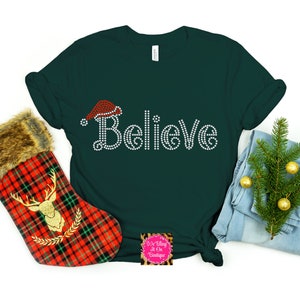 Op de afbeelding: Een donkergroen T-shirt met het woord "Believe" gespeld in witte strass steentjes. Boven het woord is een rode kerstmanmuts met witte rand. Het shirt ligt op een witte achtergrond.