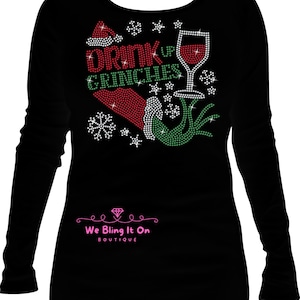 Op de afbeelding: Zwart longsleeve shirt met een strass-ontwerp met een Grinch-hand, een glas wijn, een kerstmanmuts en de tekst "Drink Up Grinches".