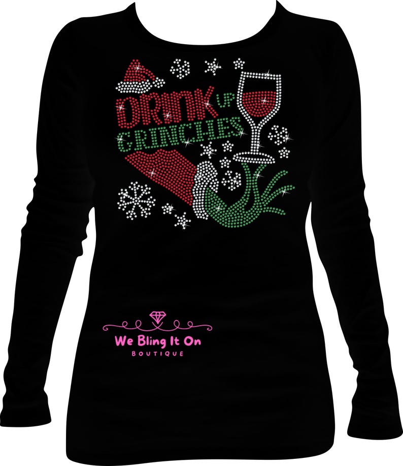Op de afbeelding: Zwart longsleeve shirt met een strass-ontwerp met een Grinch-hand, een glas wijn, een kerstmanmuts en de tekst "Drink Up Grinches".