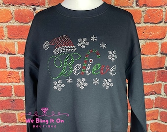 May include: Black sweatshirt with a festive design. The word "Believe" is spelled out in green and white rhinestones, accompanied by a Santa hat and snowflakes. The design is embellished with red and white rhinestones.