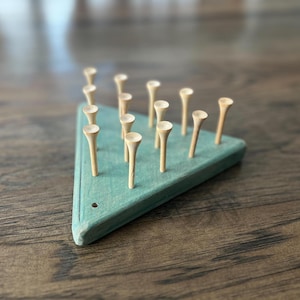 May include: A wooden triangle game board with a teal green surface and 13 wooden pegs with white tops. The pegs are arranged in a triangular pattern.