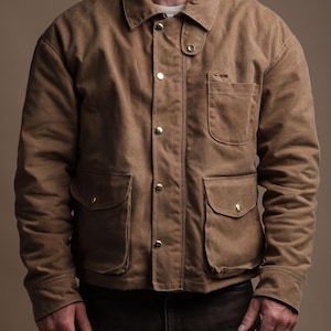 May include: A brown jacket with a collared neckline, four front pockets, and gold-colored buttons. The jacket has a rugged, workwear aesthetic. The model is wearing the jacket with dark brown pants.