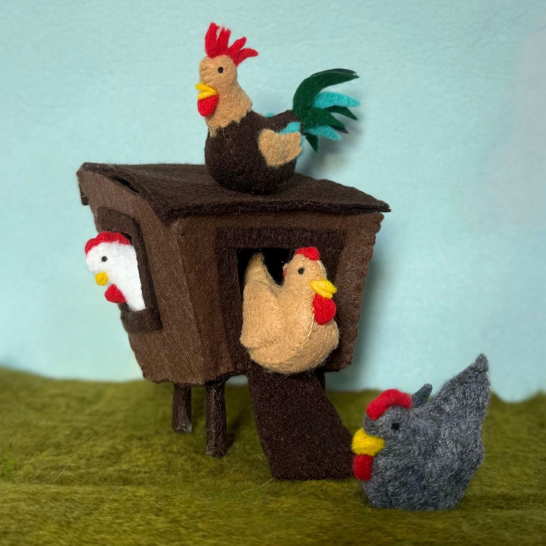 May include: A felt chicken coop with three felt chickens. The coop is brown with a chicken on top, and two chickens peeking out of the coop. A gray chicken is on the ground in front of the coop. The chickens have red combs and yellow beaks.