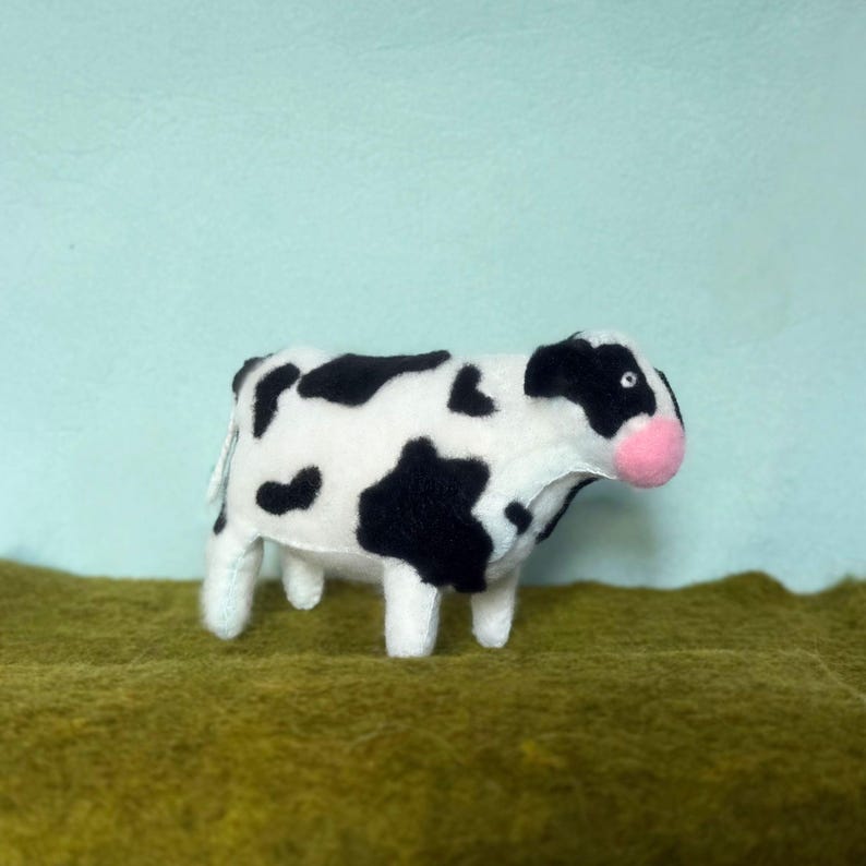 May include: A handmade stuffed cow with a black and white spotted pattern. The cow has a pink nose and is standing on a green surface against a light blue background. This plush toy is a cute farm animal.