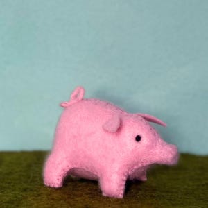 May include: A handmade pink felt pig with black eyes, a curled tail, and small ears. The pig is standing on a green surface with a light blue background. This soft toy is a charming addition to any collection.