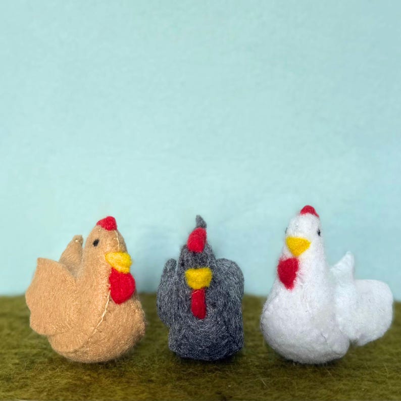 May include: Three felt chicken figurines in shades of tan, gray, and white. Each chicken has a red comb and wattle, and a yellow beak. The chickens are arranged on a green surface against a light blue background.