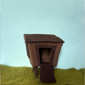 May include: A handcrafted felt house in shades of brown, with a dark brown roof and a lighter brown door. The miniature house is set against a backdrop of green and a light blue sky.
