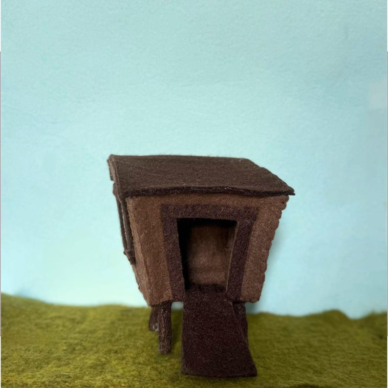 May include: A handcrafted felt house in shades of brown, with a dark brown roof and a lighter brown door. The miniature house is set against a backdrop of green and a light blue sky.